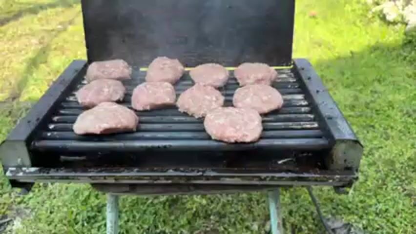 The video shows a grill sitting on a green lawn, cooking eleven raw, round, patties. Smoke is coming off the grill, which has a black top and a grey grate. The grill sits on a green stand with skinny legs. The background is blurred, but appears to be grass and a stone wall.
