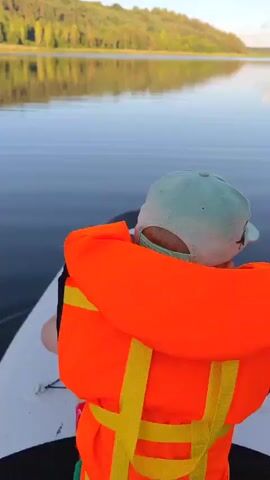 This video shows a child wearing a light blue cap and an orange life vest on a white paddleboard on a calm lake. The paddleboard has a black and white star design on its front. The child is facing away from the camera, looking towards the water and the distant shore. The lake is still, with gentle ripples, and reflects the clear blue sky and a tree-covered hillside. The sun appears to be low in the sky, casting a soft light on the landscape. The overall scene is peaceful and serene, suggesting a leisurely paddle on a summer day.