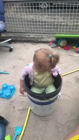 A young child is sitting inside a large white bucket with a blue and black rim in a sandbox. The child is wearing a light green t-shirt and light green pants, with a pink cardigan. The child has blonde hair pulled into a ponytail with a blue hair tie. The child is looking down and to the left. To the left of the bucket is a blue sandcastle mold. A yellow tube is partially visible in the sand to the right of the bucket. In the background, there is a metal fence and various toys. The ground is covered in sand.
