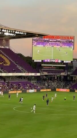 This video shows a soccer game in progress at a stadium. The scoreboard indicates the time is 00:35 and the score is 0-0. Players in white and purple uniforms are on the field, with some players actively engaged in play. Spectators are seated in the purple stadium seating. A large video screen displays a live feed of the game, as well as team logos and scores. The sky is visible in the background, with a gradient of orange and pink, suggesting it is either sunrise or sunset. The stadium is illuminated by floodlights.