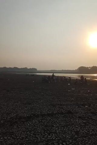 The video shows a wide shot of a dry, cracked ground with several motorcycles parked on it. In the background, there is a body of water with trees lining the far bank. The sun is setting, casting a warm glow over the scene. There are a few people visible, some sitting on the motorcycles, others standing near the water's edge. The overall impression is one of a tranquil, natural setting at the end of the day.