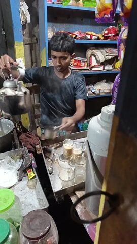 A young man is preparing drinks in a small, cluttered shop. He wears a dark t-shirt with a diamond pattern and patterned shorts. He is pouring liquid from a metal kettle into glasses arranged on a metal counter. Other items visible include jars, canisters, bags of goods, and shelves filled with packaged snacks. The environment appears to be a simple, possibly roadside, shop.