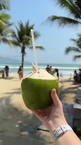 The video shows a person holding a fresh green coconut with a straw in it, against the backdrop of a sunny beach with palm trees and people in the distance. The camera then pans to an urban area with buildings and cars before focusing on a woman wearing sunglasses and a black shirt, who is walking and drinking from the coconut with the straw. She's holding the coconut in one hand while filming herself. The background shows the beach and palm trees.