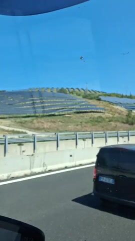 The video shows a view from a vehicle moving on a highway. On the left side of the highway, a hillside is covered with solar panels. The highway has multiple lanes and a concrete barrier separates the lanes. There are other vehicles on the highway, including cars and vans. The sky is clear and blue. The environment appears to be rural or semi-rural, with hills and vegetation visible in the distance. The video is taken from a first-person perspective, suggesting the viewer is a passenger in the vehicle.
