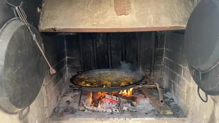 A wide shot shows a large paella pan sitting over a crackling wood fire inside a stone fireplace. The paella is a rich brown color with visible chunks of meat and vegetables, and it is bubbling and steaming. The camera slowly zooms in on the paella, offering a closer look at the ingredients and the cooking process. The fireplace is rustic, with rough stone walls and wooden beams. Two large black pots hang from hooks on either side of the fireplace opening, and a metal poker rests on the hearth. The scene is well-lit by the firelight, creating a warm and inviting atmosphere.