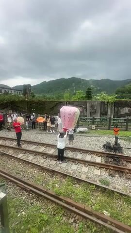 A group of people is gathered on and beside a railroad track in a somewhat rural setting. They are releasing a sky lantern, colored pink and blue, with writing on it. The lantern rises into the sky, joining several others already airborne. A backdrop of green, tree-covered hills can be seen under an overcast sky. The people are dressed casually and appear to be tourists or participants in a cultural event. The area around the tracks is covered in gravel and grass.