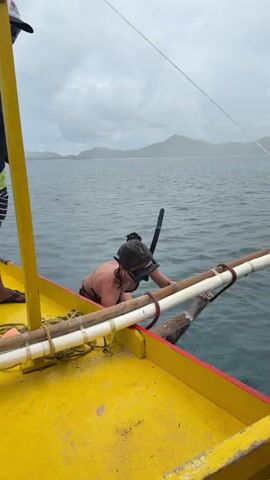 A woman in a black bikini and snorkel gear is climbing into a bright yellow boat from the ocean. The boat has white and brown bamboo-like structures attached to its side. The water is choppy and the sky is overcast. An island is visible in the background. A man's arm is partially visible on the left side of the frame. The woman is holding onto the boat's structure for support.