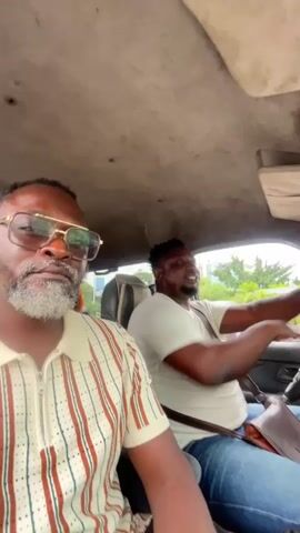 The video shows the interior of a vehicle with two men. The man in the foreground is wearing glasses and a striped collared shirt. The man in the background is driving and wearing a white t-shirt. The vehicle appears to be in motion. The interior of the vehicle is visible, as is some of the environment outside through the windows.