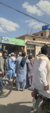 The video depicts a bustling street scene in Kuchlak, Pakistan, during the daytime under a clear, slightly cloudy sky. A large number of men, predominantly wearing traditional shalwar kameez, are gathered in what appears to be a commercial or market area. Some children are also visible in the crowd.

In the foreground and midground, various individuals are standing, talking, and gesturing. Two men, in particular, stand out due to their attire: one wears a black shalwar kameez, a black vest, and carries a rifle, while another man in similar tactical-style gear is also seen. Their presence suggests a security detail or an armed group within the crowd. An individual in a light blue shalwar kameez and a man pushing a handcart are also visible.

The background features several single and multi-story buildings, some with shops on the ground floor. Visible signs include a green and white one reading "عبداللہ جنرل سٹور" (Abdullah General Store) and a blue and yellow sign with "افغان میڈیکل جنرل سٹور" (Afghan Medical General Store) and "افغان" (Afghan) written on it, indicating local businesses. Overhead, power lines crisscross the street. To the right, part of a construction vehicle, likely an excavator or backhoe, with "HYUNDAI" visible on its arm, is parked or in operation within the crowded area. A blue water tank can be seen on the rooftop of one of the buildings.

The overall impression is of a lively street with a significant public presence, possibly during a public gathering, procession, or a busy market day, amplified by the presence of armed individuals.