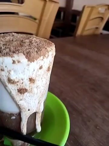 Here is a description of the video.
The video shows a glass of coffee on a green saucer on a wooden table. The coffee has a layer of what appears to be white foam on top, with chocolate powder sprinkled on it. Some of the foam has dripped down the side of the glass. A black straw and a silver teaspoon are placed beside the glass on the saucer. There are two chairs visible in the background, and music is playing.
