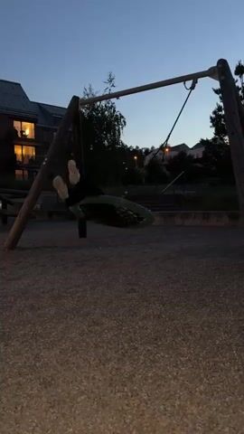 A person is seen swinging on a tire swing in a playground at dusk. The swing set is made of wood. The ground is covered in gravel. In the background, there is a building with lights on, trees, and a suburban hillside. The sky is dark blue. The person on the swing is wearing dark pants and a white jacket. The swing is circular and made of a green material. The person's movements are blurred due to the motion of the swing.