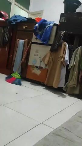 A hand enters the frame from the right and makes a waving gesture. The scene behind the hand is an indoor space with tiled flooring and cluttered furniture. A brown wooden cabinet is on the left, draped with various garments and fabrics. Next to it is a tall, beige appliance, possibly a refrigerator, with graphics on its front. More clothing hangs on a rack to the right of the appliance. A broom with colorful bristles rests on the floor in the lower left corner. The lighting appears to be artificial, creating a somewhat dim atmosphere.