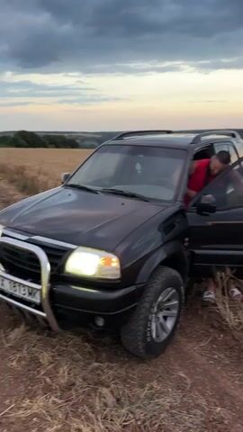 A black SUV is parked off-road in a field of dry, golden grass or wheat. The driver's side door is open, and a person wearing a red shirt is visible inside the vehicle. The camera pans away from the SUV to show a wide landscape with rolling hills and a cloudy sky at dusk, with hints of orange and pink in the horizon. The SUV is seen again as the camera continues to pan across the field, eventually focusing on a dirt path leading towards a lone tree in the distance.
