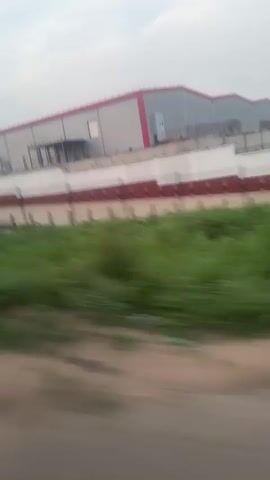The camera captures a moving view of a large industrial building. The building has a light gray exterior with a red stripe along the roofline. In front of the building, there's a striped barrier with a repeating pattern of white and dark red sections. A grassy area stretches between the road and the barrier. The ground beside the road is dirt. The sky is overcast, and the overall lighting is muted. The camera's motion creates a blur effect, particularly on the foreground elements.
