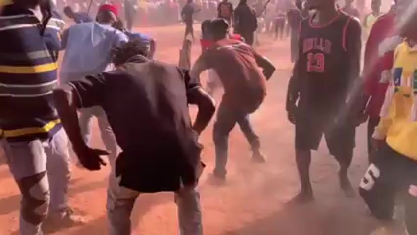 The video depicts a densely populated outdoor scene, likely a gathering or celebration, taking place on a reddish-brown dirt surface. Numerous people are visible, most of whom appear to be African. They are dressed in various casual clothing. The environment is dusty, with the dust seemingly kicked up by the activity of the crowd. In the background are several huts or small buildings with tin roofs, suggesting a rural setting. Some individuals are interacting and moving around, with several people capturing the event on their mobile phones. The lighting suggests it is daytime.