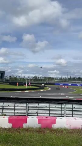The video shows a go-kart race on a cloudy day. The camera is positioned behind a barrier of pink and white plastic blocks and green grass, looking out over a black asphalt race track with red and white curbing. In the distance, there are several go-karts racing. One go-kart, colored pink, can be seen on the left side of the track turning a corner. Further down the track, two other go-karts are visible, with one turning and the other heading straight. Beyond the track, there are buildings, trees, and lampposts. The sky is filled with large, white cumulus clouds against a pale blue sky.