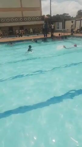 The video shows a swimming pool with several people in and around it. In the pool, individuals are swimming, with one child visible in the foreground, doggy-paddling towards the center. Other swimmers are visible further in the background. Surrounding the pool are people standing or sitting, some appearing to be watching the swimmers. A building is also visible in the background, with a pattern on its facade. The pool water is light blue and there are shadows visible at the bottom of the pool.