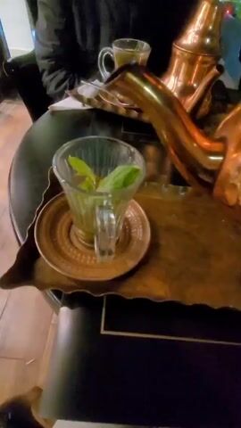 A person is pouring tea from a brass teapot into a clear glass cup filled with mint leaves. The cup sits on a saucer on a brass tray. Another cup of tea is on the table in the background next to the teapot. The person pouring is wearing a dark jacket. The table is round and black. The floor is wooden.