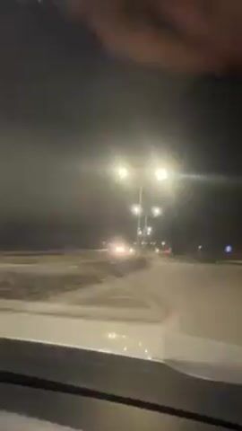 A car is driving down a road at night. The road has multiple lanes and bright streetlights. A few cars are driving in the opposite direction. A hand briefly appears in the frame at the top. The rest of the video is shot from the perspective of the driver.