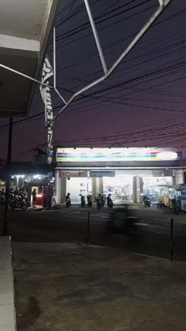 This media file captures a street scene at dusk in Jember, Indonesia. The dominant subject is a brightly lit convenience store, likely a branch of a chain with a prominent white sign displaying a multicolored logo. In front of the store, several motorcycles are parked, and a few people are visible, though their features are indistinct due to the low light and motion blur. Across the street, more vehicles, including what appears to be a yellow food cart, are present.

The sky is a deep purple, transitioning to lighter shades of pink and orange near the horizon, indicating sunset or twilight. The overhead power lines are a prominent feature, crisscrossing the frame. The overall atmosphere is one of a quiet evening in a semi-urban or commercial area. There are no strong emotional cues, but the lighting and setting suggest a typical end-of-day scene. No other significant text is clearly legible in the media, apart from the store's branding.