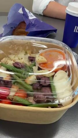 A hand is shown lifting a clear plastic lid off a takeout container filled with a salad. The salad contains green beans, cherry tomatoes, olives, cubed potatoes, pieces of bread, and a small container of orange dressing. A hard-boiled egg is also visible on the side. In the background, a blue paper bag with a croissant inside and a blue paper cup with the logo "IL" are on a grey surface. The video is shot from a close-up perspective, focusing on the food.