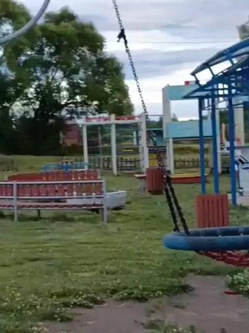 This is a video of an outdoor playground. The playground has swings, a sandbox, and some exercise equipment. The weather appears to be overcast, and the time of day is likely daytime. The video is set in a grassy area with trees in the background. There are no people visible in the video until the very end, where a person is seen in the distance interacting with a swing set. There is no visible text in the video.