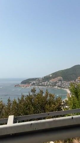 A panoramic view captures a coastal city nestled against a lush green hill under a clear blue sky. The azure sea is dotted with numerous boats, some appearing to be speedboats leaving white wakes, while others are moored in a harbor. A section of the coastline features a sandy beach lined with white umbrellas and lounge chairs, indicating a popular tourist destination. In the foreground, a metal railing runs horizontally, suggesting the video was filmed from an elevated vantage point, possibly a balcony or a viewpoint overlooking the bay. Vegetation, including trees and shrubs, is visible in the foreground and on the hillside. The overall scene conveys a bright, sunny day at a seaside resort.