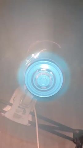 A close-up, downward-facing view shows a blue fan with concentric rings on its grille. The fan blades are spinning, creating a blur of light and air. The fan is mounted on a white articulated arm with a black base. A white electrical cord hangs down from the fan. The background is a muted, dusty blue. The overall impression is one of a functional, but perhaps aging, piece of equipment.