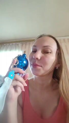 A woman with long blonde hair, wearing a pink tank top, holds a blue bear-shaped fan with a white lanyard to her face. She appears to be speaking and possibly fanning herself, her eyes are closed in the first frame and open in the next. The background consists of a wall with a curtain and a dresser.