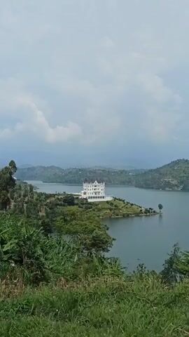 A long shot shows a large, white, ornately-designed building in the middle of a lake, from an elevated position with thick green foliage at the very bottom of the frame. The large building, which may be a castle, has dark conical peaks. The sky is overcast with gray clouds. The water is calm. Hills are visible in the background behind the lake. There is a small patch of green grass surrounding the building.