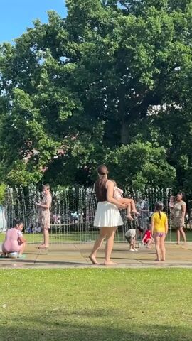 The video captures a sunny day at a public park with a fountain. Several children and adults are present, some interacting with the fountain. In the foreground, a woman in a white skirt and brown top is holding a child. To her left, a person in a pink outfit is crouched near the fountain's edge. Other children are scattered around the fountain, with some appearing to play in the water. A large, leafy tree dominates the background, with buildings and more trees visible beyond. The overall scene suggests a recreational setting on a warm day.