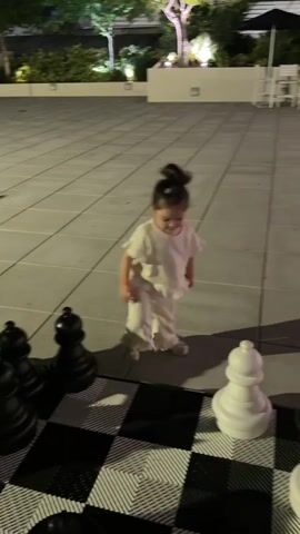 A young child in a white outfit stands on a large outdoor chessboard. The child is blurred due to movement. Large black and white chess pieces are visible on the board. The background shows a paved area with some scattered chairs and a table, along with some greenery and lighting.