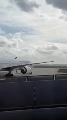 The video shows an Air France plane on a runway. The plane is white with the Air France logo on its side and the French flag on its tail. A smaller vehicle drives on the ramp behind the plane. The sky is cloudy. The camera is behind a fence, likely at an airport. The audio describes the plane is arriving at Terminal 2E, Gate L.