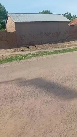 The video shows a dirt road in what appears to be a rural or semi-rural setting. On the left side of the road, there are buildings with mud or stucco walls and corrugated metal roofs. Some of these buildings appear to be homes or shops, with one structure featuring a thatched roof. A tall green and white pole stands near the road. Further down the road, a person wearing a long, light-colored garment and a head covering walks from right to left across the frame. A red motorcycle is parked next to the pole. On the right side of the road, there is a building that looks like a mosque, with a white minaret topped by a green dome. A large tree with green foliage is also visible on this side. The sky is clear and blue, suggesting a sunny day. The road surface is dry and dusty.