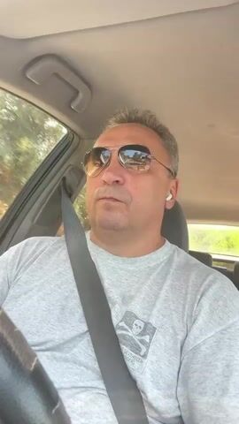 A middle-aged man with short gray hair and sunglasses is driving a car. He is wearing a gray t-shirt with a skull and crossbones graphic. He has a seatbelt on. He is wearing white wireless earbuds. The car appears to be moving on a road with trees and greenery visible outside the window. The sun is shining.