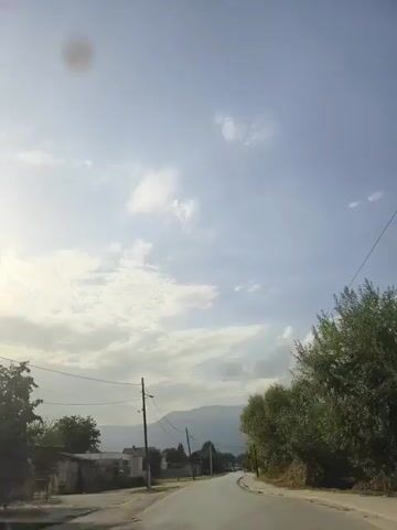 This is a first-person view video taken from a vehicle traveling on a paved road through a rural or suburban area. The sky is mostly cloudy with patches of blue visible. There are power lines with utility poles running along the side of the road. On the left side of the road, there are some buildings, including what appears to be a small house and some agricultural structures. On the right side of the road, there are trees and vegetation. In the distance, mountains are visible. The road curves to the right as the vehicle continues to move forward.