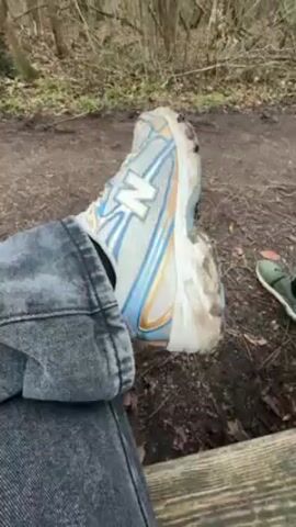 The video shows a person sitting on a bench in a forest or park. The camera focuses on the person's dirty New Balance shoe, which is light blue with a white "N" logo. The person is wearing dark gray jeans. The ground is covered in dirt and leaves, and there are trees in the background. The other shoe is green. The scene is overcast.