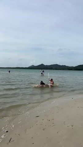 Two individuals are in a transparent kayak near a sandy beach. The person in the front is seated, facing forward, while the person in the back is also seated and holding a paddle, actively paddling the kayak. The water is relatively calm with small waves breaking on the shore. In the background, several other people are standing in the water further from the shore, and a distant coastline with hills is visible under a cloudy sky. The scene depicts recreational activity on a body of water adjacent to a beach.