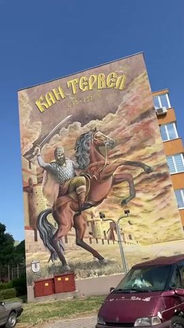 The video pans across a large mural painted on the side of a building. The mural depicts a historical figure on horseback, wielding a sword. The figure appears to be a warrior, dressed in armor and a helmet, with a beard. The horse is rearing up, adding to the dramatic depiction. In the background of the mural, there are fortifications that resemble a castle or a walled city. The sky in the mural is depicted with dramatic clouds and warm, fiery colors, suggesting a battle or a significant event. The text above the figure appears to be in Cyrillic script and reads "КАН ТЕРВЕЛ" which translates to "Khan Tervel." Below this, there are dates "685-721". The building itself is a multi-story structure, likely an apartment building, with windows visible. In the foreground, there is a parked red van and a portion of a silver car. The environment appears to be an urban or suburban setting, with some greenery visible to the left of the building and a clear blue sky above. The sun is shining, casting shadows.