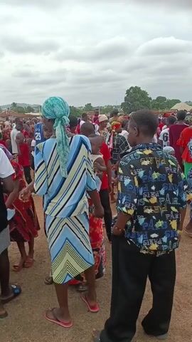 The video depicts a crowd of people gathered in an outdoor space with a dirt ground. Most are dressed in casual clothing, and there are several children in the crowd.  Some individuals are wearing traditional African attire, and head coverings are common.  The sky is overcast. The focus shifts from a woman in a blue and yellow patterned dress and headscarf to various individuals within the crowd, offering a glimpse of their clothing and interactions. The environment seems to be a village or community gathering space, with residential buildings visible in the background.