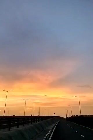 A low-angle video, shot from a moving vehicle, captures a sunset over a highway. The sky is a gradient of deep blue to orange and yellow as the sun sets behind scattered clouds. Streetlights line the sides of the elevated highway, some illuminated. The road ahead curves gently to the right, with a dark tree line visible in the distance. The protective barriers of the highway are visible on either side of the asphalt road.