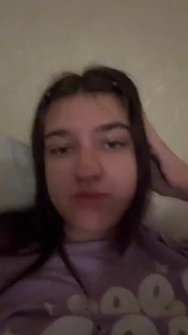 A medium shot shows a young woman with dark hair lying down and looking at the camera. She is wearing a purple t-shirt with white graphics. She has her right hand on the back of her head. The background appears to be a wall with a faint textured pattern.