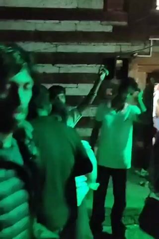 THE VIDEO SHOWS A GROUP OF PEOPLE DANCING UNDER GREEN LIGHTS. THE SCENE APPEARS TO BE AN OUTDOOR GATHERING, POSSIBLY A CELEBRATION OR PARTY. THE BACKGROUND CONSISTS OF BRICK WALLS AND DOORS, SUGGESTING A RESIDENTIAL OR URBAN ENVIRONMENT. PEOPLE ARE IN MOTION, WITH SOME RAISING THEIR ARMS AND MOVING TO THE MUSIC. THE OVERALL ATMOSPHERE IS LIVELY AND FESTIVE.
