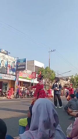 A group of people are sitting on the ground watching a street performance. In the center of the street, a dancer in a vibrant red and gold costume is performing a traditional dance. She has her arms outstretched and is wearing an elaborate headdress. Behind her, other dancers in similar attire are also visible. A woman in a black jacket and hijab stands to the side, watching the performance and holding a beverage. The street is lined with shops and buildings adorned with advertisements. Several motorcycles are parked along the side of the road. The sky is clear and blue, indicating a sunny day. The spectators in the foreground are mostly women, some wearing headscarves. One person is wearing jeans and a black t-shirt. The overall atmosphere appears to be festive and engaging.