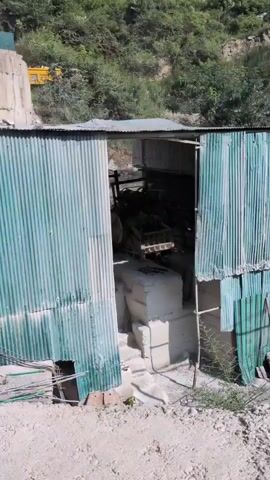 A teal excavator is actively demolishing a concrete structure. It is positioned on top of the structure and is using its bucket to break apart the concrete. A yellow vehicle, possibly a truck, is visible in the background, further up the hillside. Below the demolished structure is a corrugated metal building with a green-tinted roof and walls. The building appears to be open on one side, revealing machinery and stacked materials inside. The environment is a hilly, vegetated area, with trees and foliage covering the slopes. The ground in the foreground is dirt and gravel. The overall scene suggests construction or demolition activity in a somewhat industrial setting within a natural landscape.