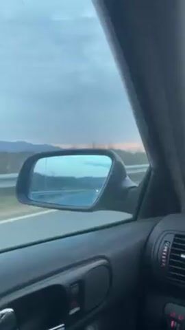 The camera is placed inside a car that is traveling down a highway. The view is from the passenger side, focusing on the side mirror. The mirror reflects the highway stretching out into the distance, flanked by fields and hills. The sky is overcast with a hint of orange at the horizon, indicating either sunrise or sunset. The car's interior, including the door panel, window controls, and dashboard air vents, are visible. The car appears to be moving at a steady speed.