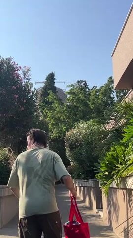 A person walks away from the camera down a paved path. They are wearing a light green t-shirt and dark pants, and they are carrying a red bag in their right hand. The t-shirt appears to be wet with sweat. The path is bordered by greenery on both sides, with trees, bushes, and flowering plants visible. A building with a crane is visible in the background. The sky is clear and blue. The lighting suggests it is daytime.