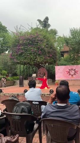 A woman in a red and gold dress is performing a traditional dance on a red brick stage. Behind her is a large metal sculpture and a banner with a pink floral design. Several people sit in folding chairs watching her performance. The setting is an outdoor courtyard, with green trees and purple flowers.