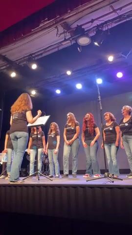 A choir of women stands on a stage, singing. A woman with her back to the camera, wearing a black t-shirt and blue jeans, appears to be leading the choir, holding a music stand. The choir members, also wearing black t-shirts and blue jeans, are arranged in two rows. They are all looking towards the leader and singing. The stage is lit with stage lights, and a red curtain is visible at the top of the frame.