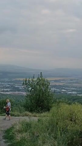 The video opens with a wide shot of a landscape from a high vantage point. The sky is overcast with gray clouds. In the foreground, two people are visible. A woman in white pants and a white top is walking away from the camera. A man in a gray shirt and dark shorts is standing facing away from the camera, holding what appears to be a camera or bag. They are on a dirt path. Behind them, a valley stretches out, dotted with buildings and trees. In the distance, rolling hills and mountains can be seen under the hazy sky. The camera pans slowly to the right, revealing more of the valley and the distant hills. The vegetation in the foreground consists of green grass and some white wildflowers. The video transitions to shots of the landscape without the people, showing different sections of the valley and the distant mountain ranges. The overall impression is of a panoramic view of a rural or semi-rural area under a cloudy sky, possibly at dusk.